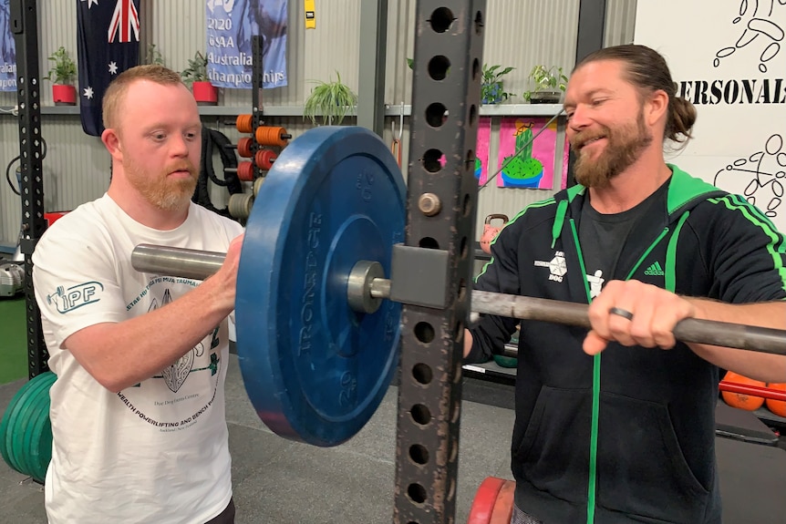 Jack Carroll adjust a weight on a barbell in a gym with trainer Shannon Pigdon looking on.