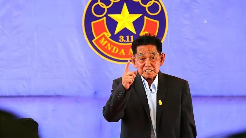 Peng Jiasheng speaks with one finger raised in front of the logo of the MNDA. 