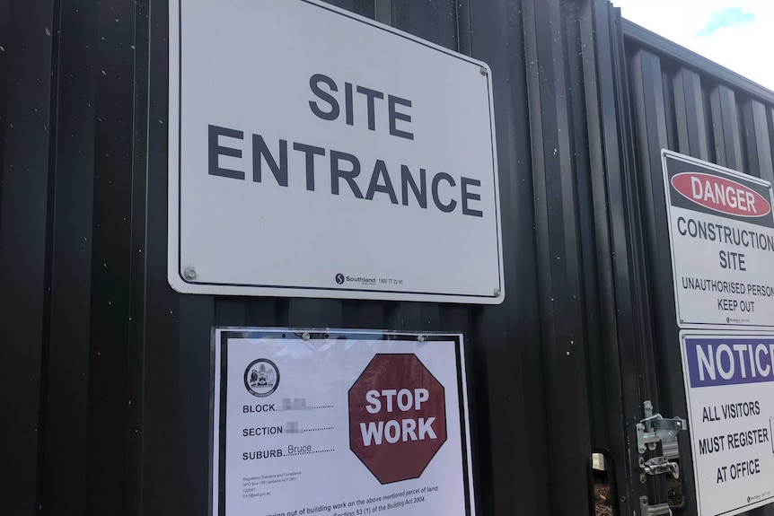 Closeup of a notice with a red stop work sign below a white and black site entrance sign on a black fence.