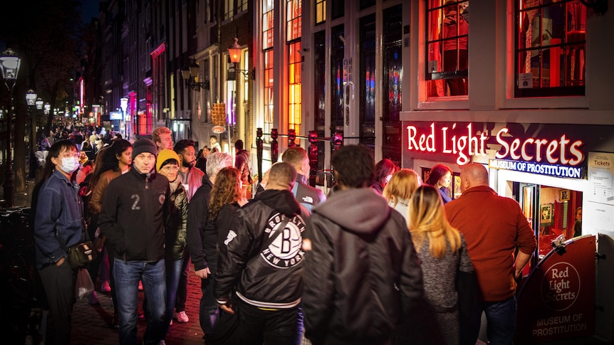 A large crowd of people walking through a street. On the right side building fronts and a sign that says 'Red Light Secrets'.
