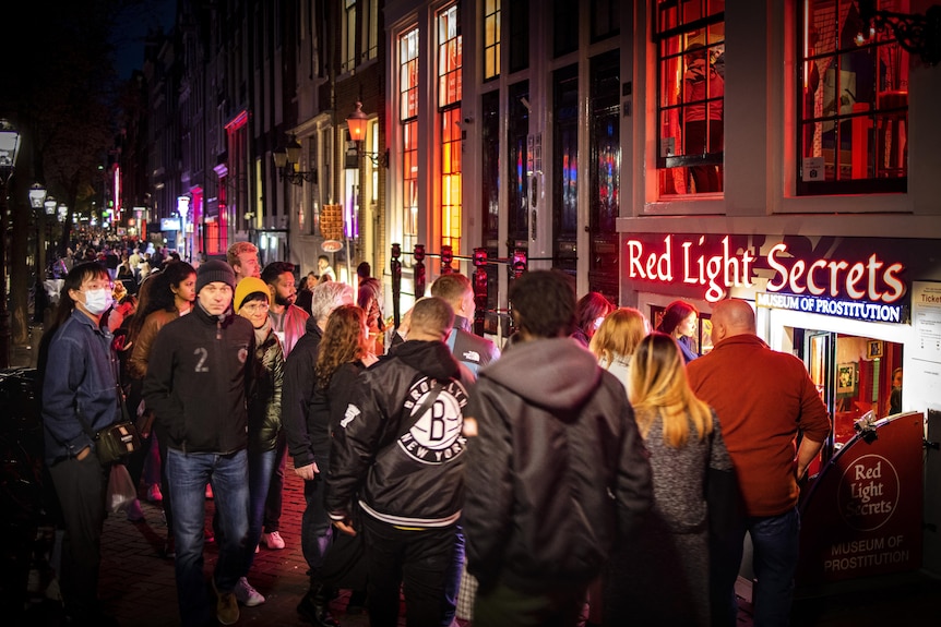 A large crowd of people walking through a street. On the right side building fronts and a sign that says 'Red Light Secrets'.