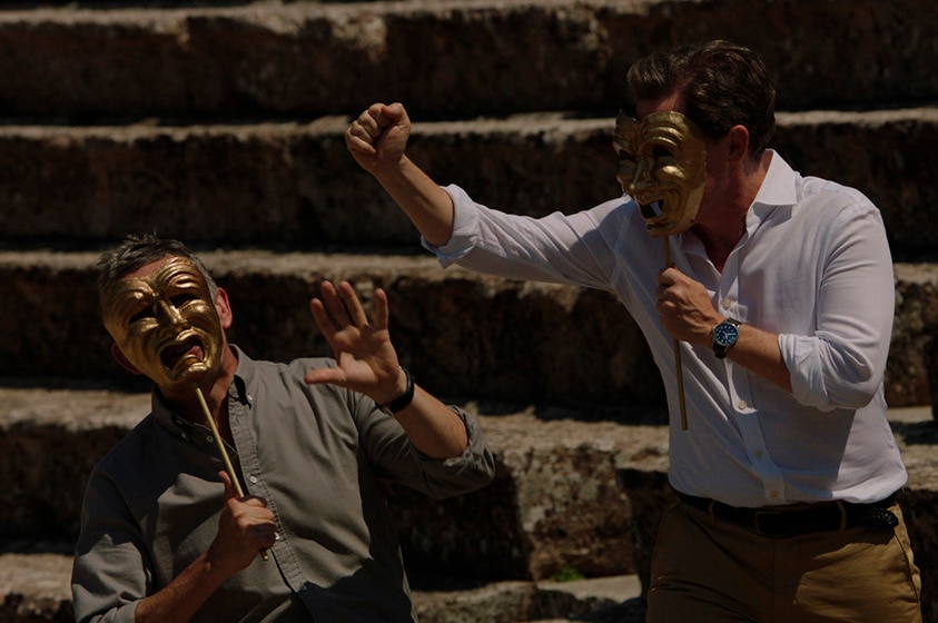 Two men wear button up shirts and pose with gold Greek theatre masks in front of stone step ruins.
