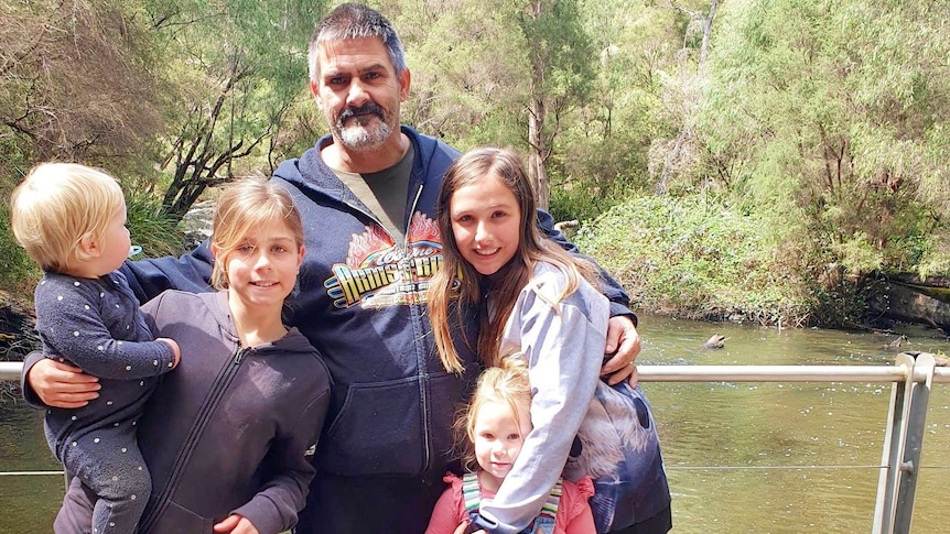 A man stands in front of a creek and bushland with his four children