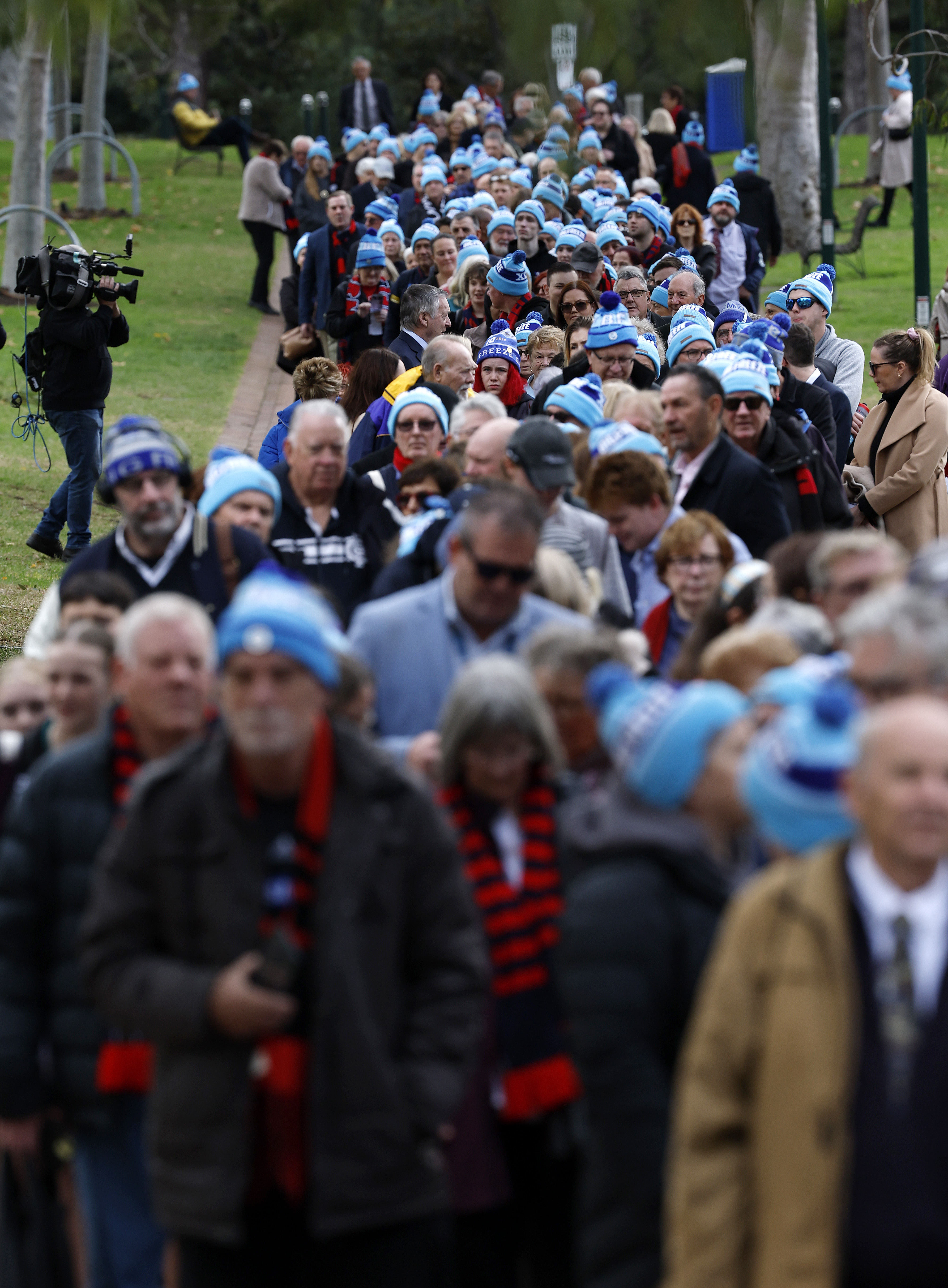 A large line of people stretches back to the rear of the shot, with most people wearing a sky blue beanie.