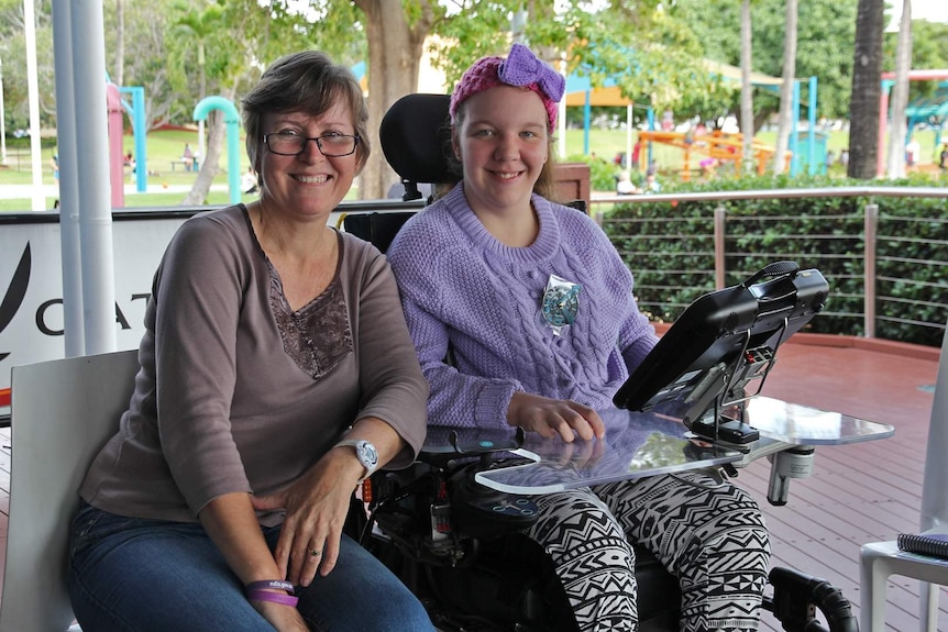 A woman in glasses sits next to her daughter who is in a wheelchair and has a tablet attached to her chair