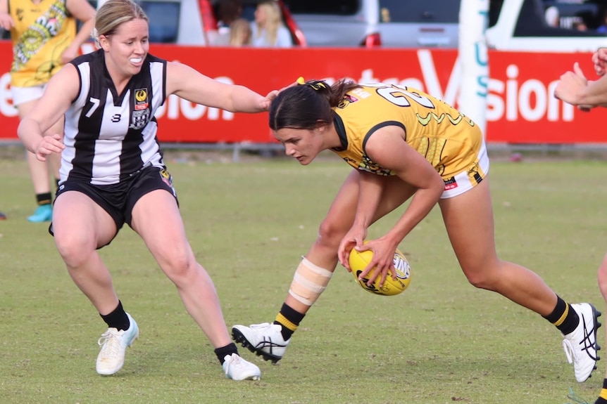 A women in black and white uniform attempts to tackle a woman wearing a yellow uniform and holding a football.