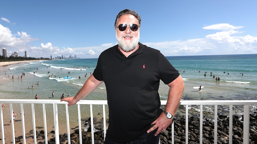 russell crowe in a black polo and shades leans on a guard rail in front of burleigh heads beach