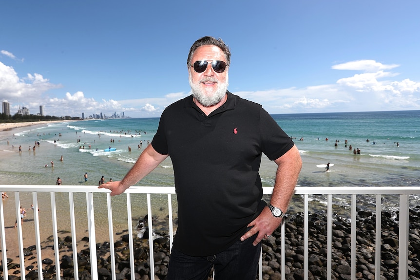 russell crowe in a black polo and shades leans on a guard rail in front of burleigh heads beach