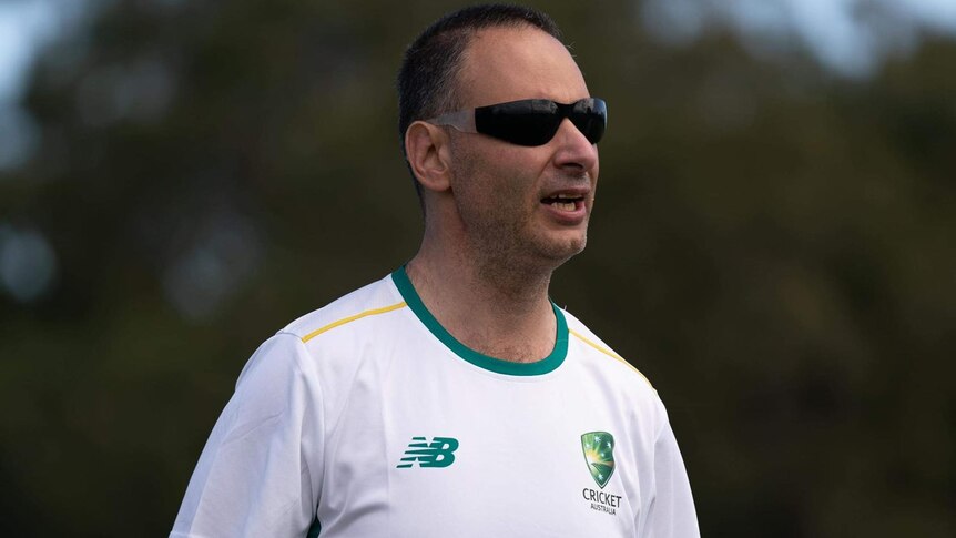 man with dark sunglasses on and white cricket t-shirt with green trim holding cricket bat