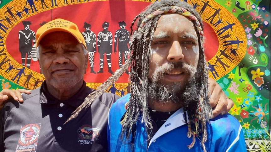 Two Indigenous man stand together with their arms around each other in front of a colourful banner.