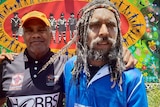 Two Indigenous man stand together with their arms around each other in front of a colourful banner.