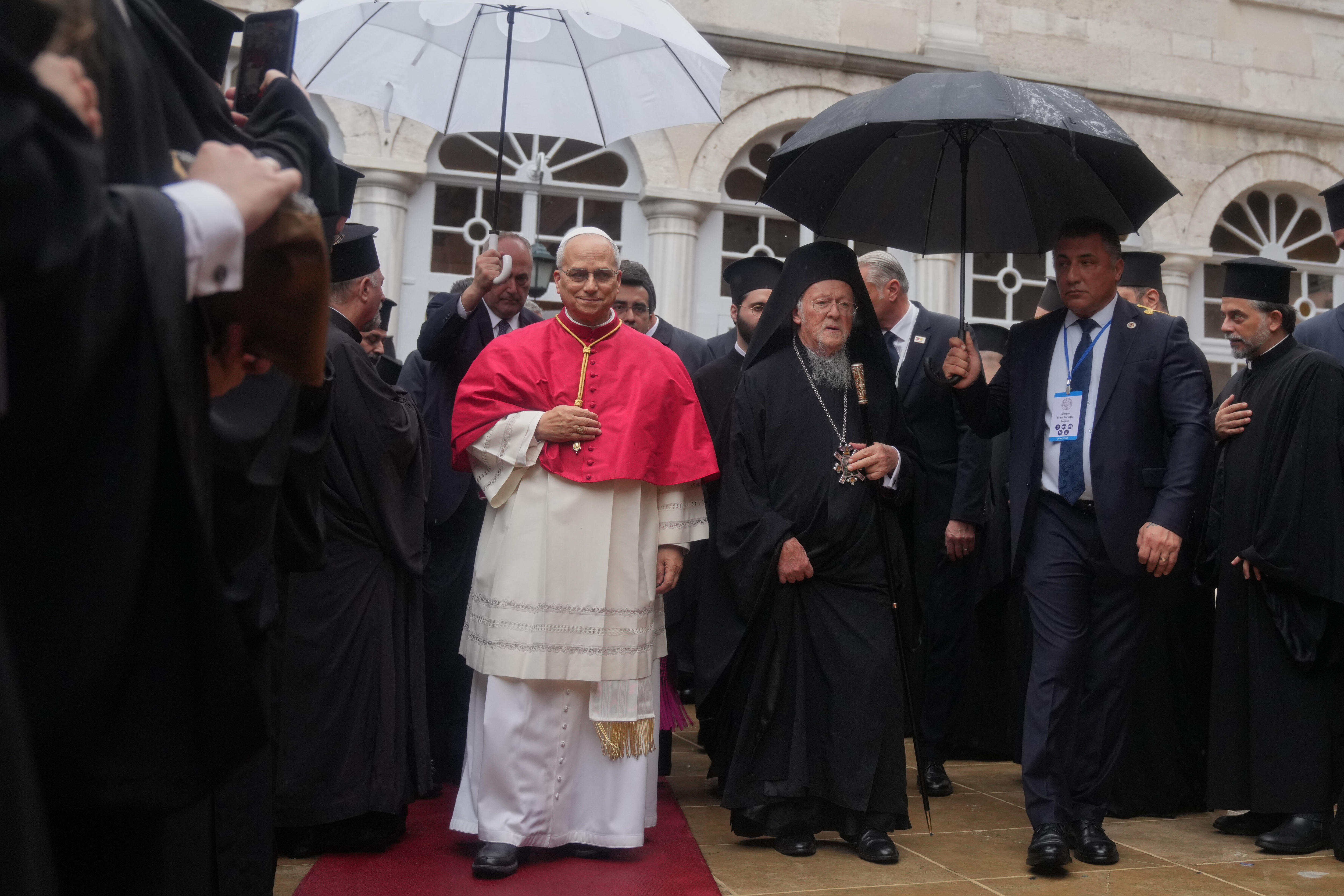 El Papa León camina con una túnica blanca y roja, y junto a él hay personas con paraguas. 