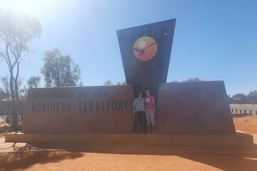 Two women stand in front of a monument that says "Welcome to the Northern Territory".
