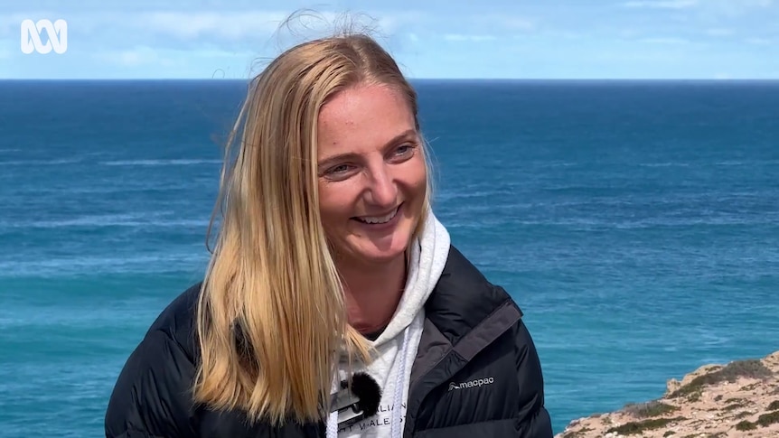 A smiling woman with blonde hair and a microphone clipped to her jumper stands with the ocean behind her