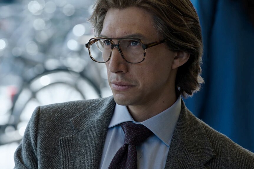 Man with glasses, greying hair and textured grey suit over a pale blue shirt looks offscreen with intensity.