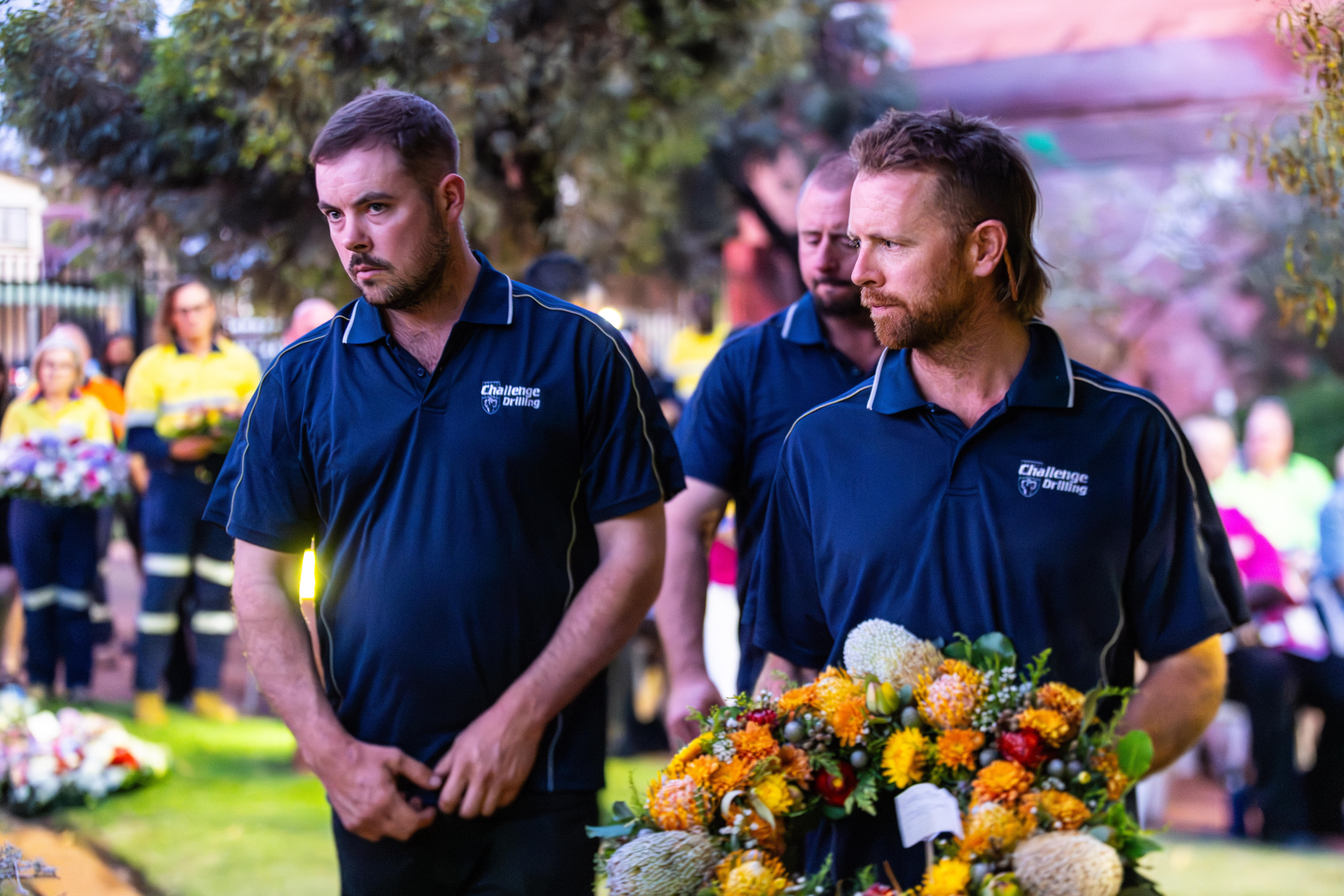 Tres hombres en un servicio conmemorativo, uno de ellos sosteniendo una corona de flores.  