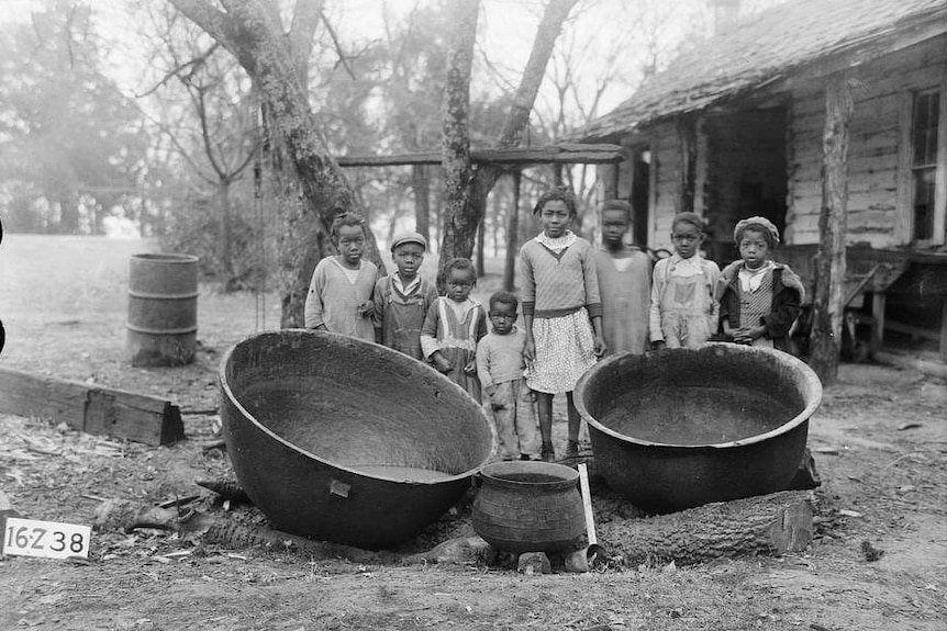 You view a black and white image of African American children in front of a derelict wooden house.