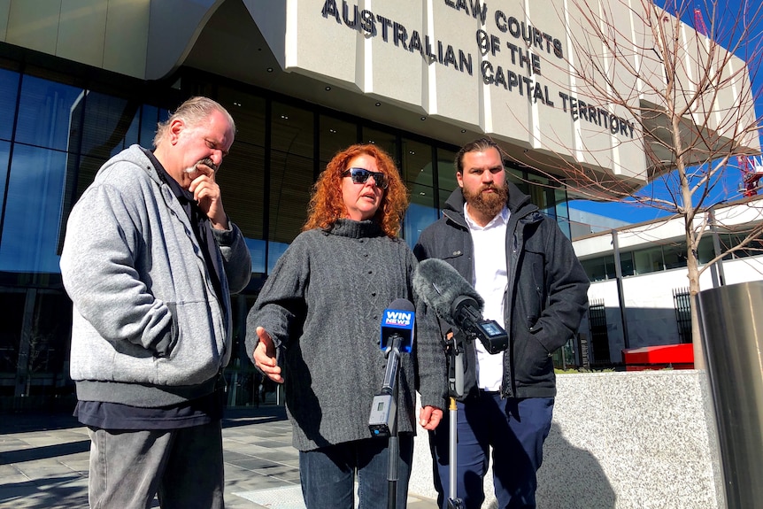 A man with a moustache, woman with orange hair and bearded man stands out the front of the ACT Law Courts. 