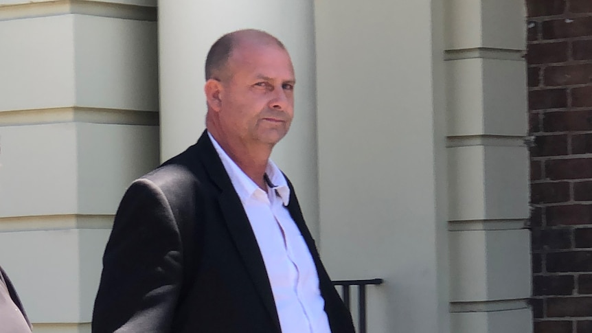 Todd Barry Apted looks right, towards the camera as he leaves a court house. He is expressionless, apart from a side-eye.