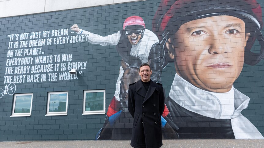A jockey clad in black stands in front of a wall featuring a mural of him standing in silks and saluting the crowd.