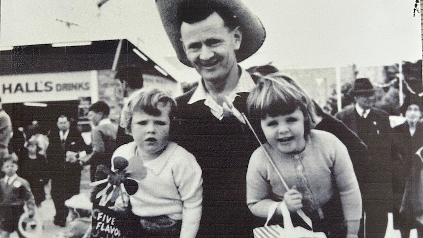 balc & white image of a man in a cowboy hat holding girls under his arms holding showbags