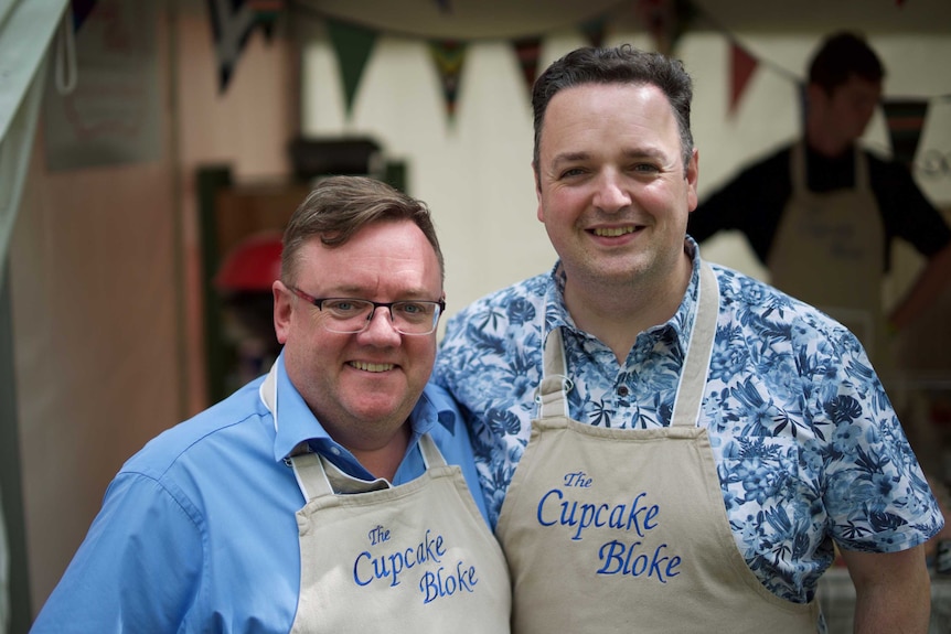 Couple Graham Herterich and Daithi Kelleher stand in Cupcake Bloke aprons, hugging and smiling.