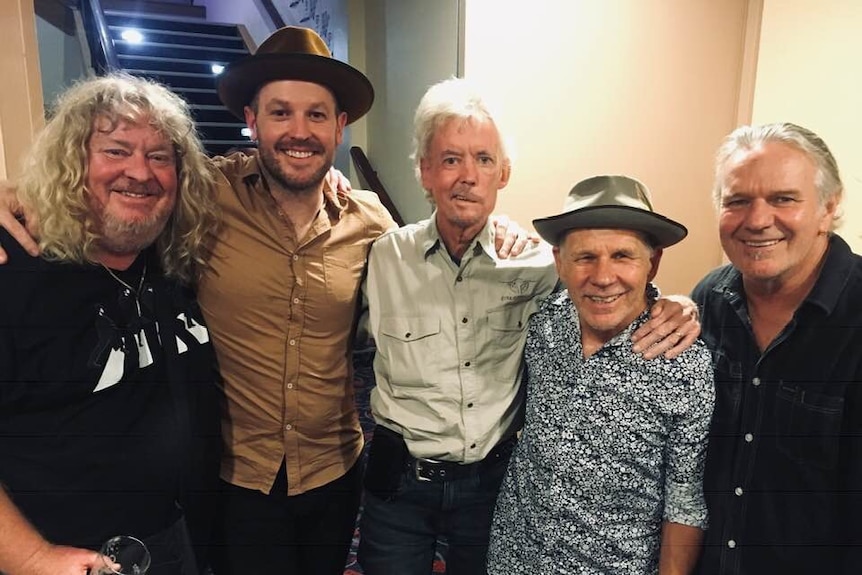 Five men stand arm in arm backstage at a music concert.