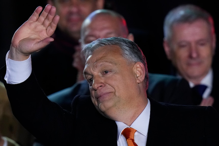 Hungary's Prime Minister Viktor Orban wears a suit and waves, standing in front of other men in suits.
