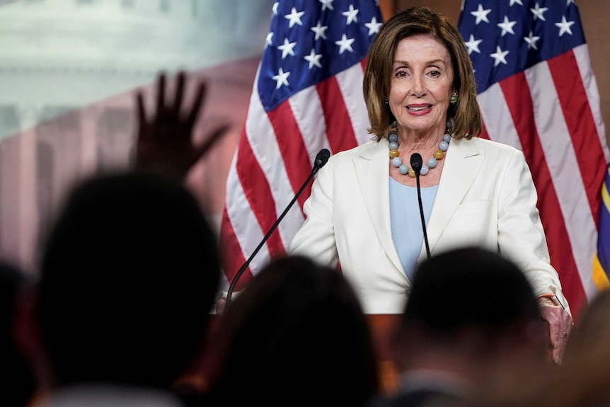 A woman in a white suit stands on a stage flanked by US flags