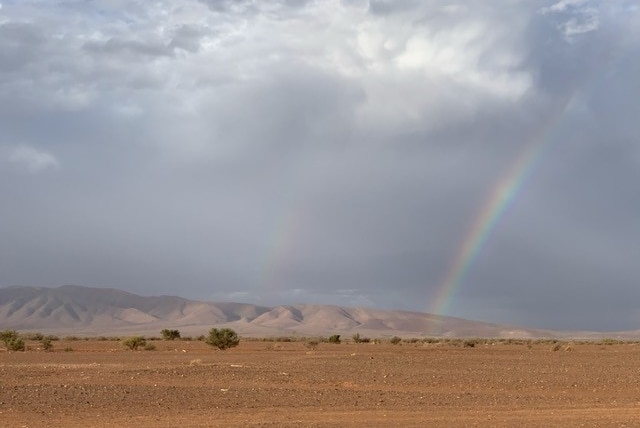 A property with red dirt, mountains in the background, and a rainbow rising out from the horizon.