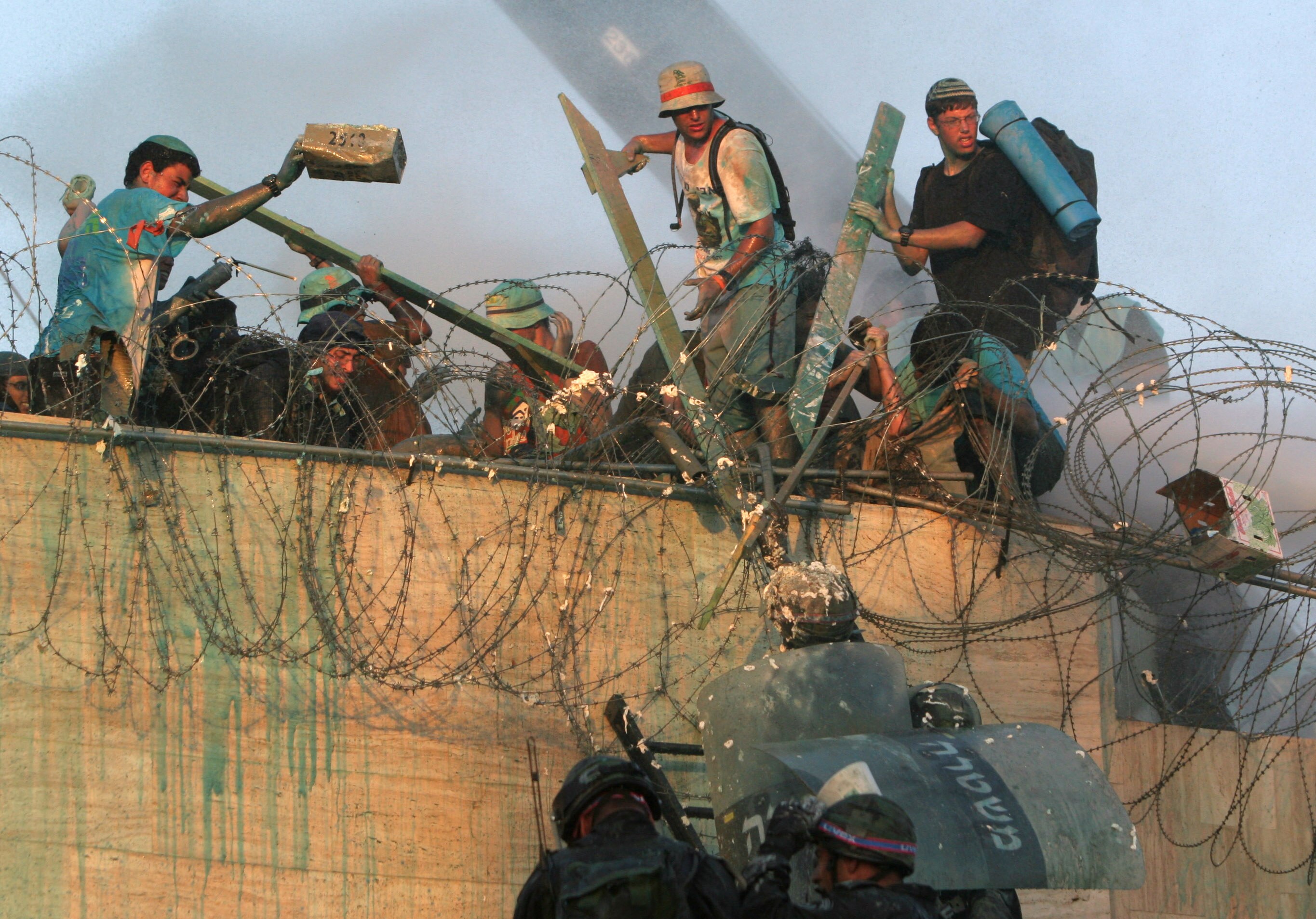 Political activists wearing hats and kippahs standing on a concrete fence using palings to push away Israeli police shields