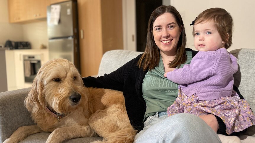 A woman sits on a couch smiling and holding a little girl, with a dog beside them.