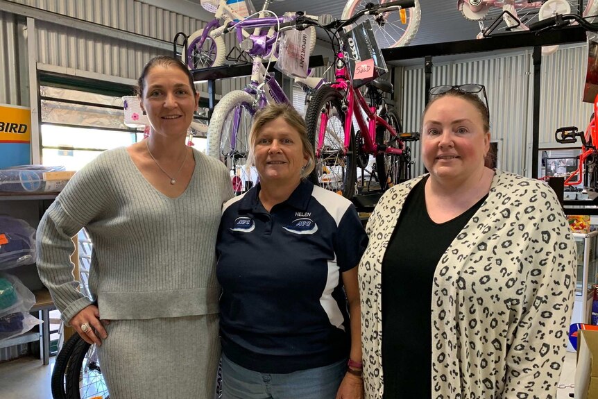 Three women stand in front of a bike rack, with bikes in a tin shed shop.