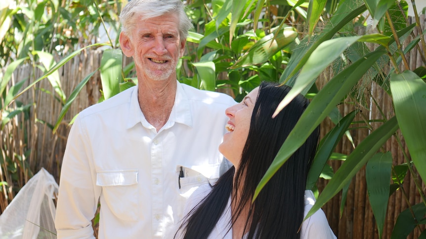 Laughing young woman, long black hair, looks back at smiling older man, grey hair and beard. Both wear white, stand in garden.