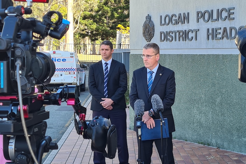 Two police detectives stand in front of a tv news cameras.