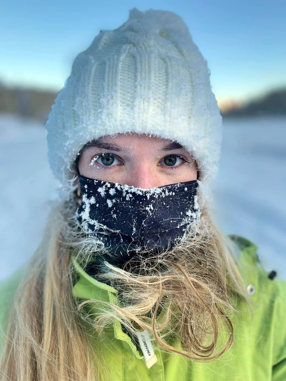 Woman with blond hear is wearing a white beanie and black face mask and has icicles on her mask and eyelashes.