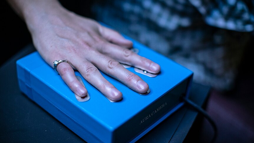 A hand with a ring with a jewel on the little finger rests on top of a blue box with metal plates in the shape of a hand.