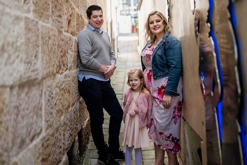 A woman stands in a laneway in Sydney with her husband and young daughter. They're all smiling.