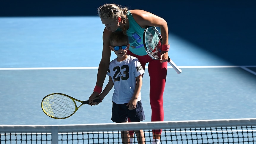 A female tennis player helps her son hold a racquet on the court.