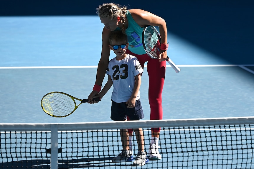 A female tennis player helps her son hold a racquet on the court.