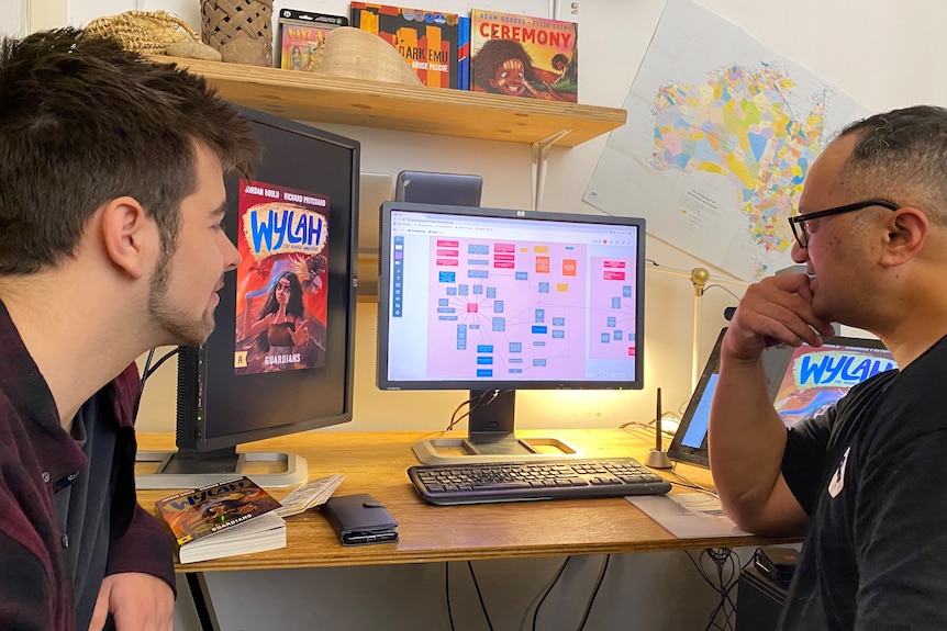 two men look at a computer screen in a room surrounded by aboriginal posters and books