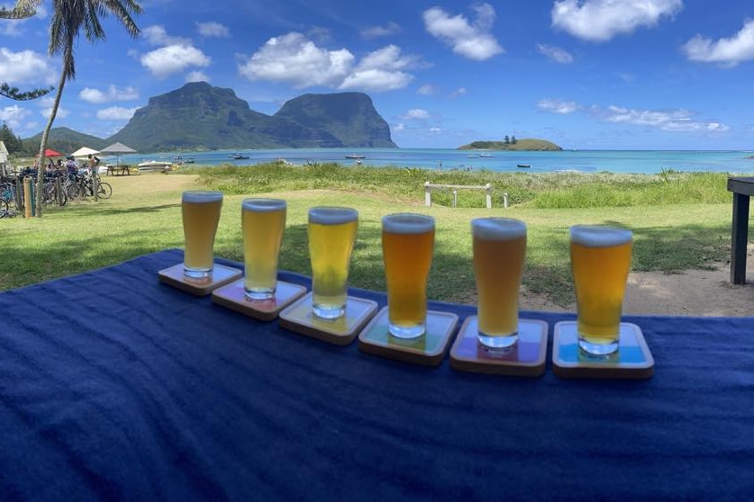 Six beers lined up on a table with Lord Howe Island waterfront in the background. 
