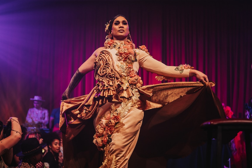 A performer walks across the stage in a floral gown, basked in golden light.