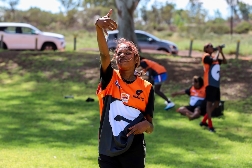 Young Aboriginal woman throws hand sign while wearing brightly coloured football jumper on football ground 
