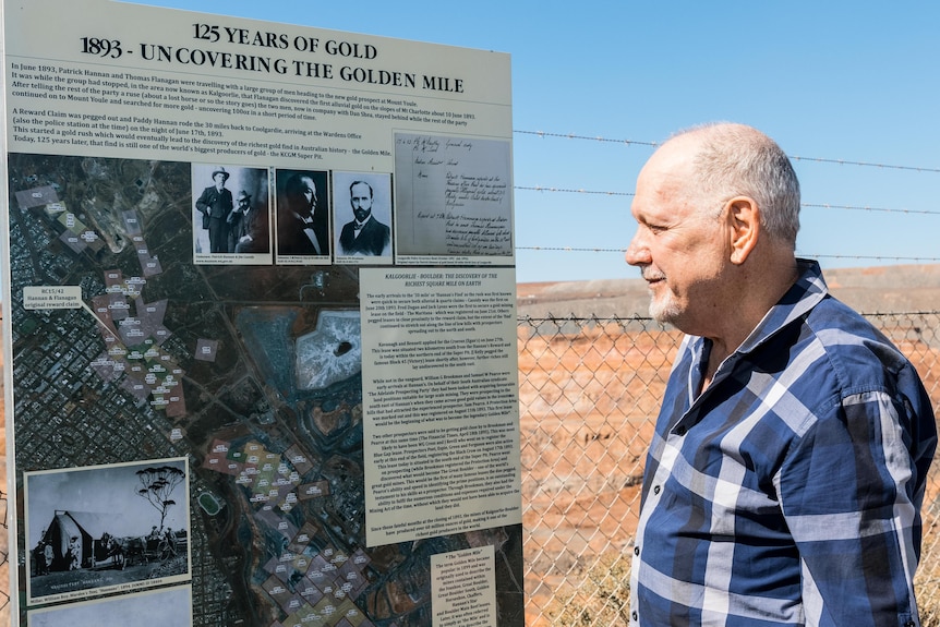 A man standing next to a public information sign at a public lookout overlooking a gold mine. 
