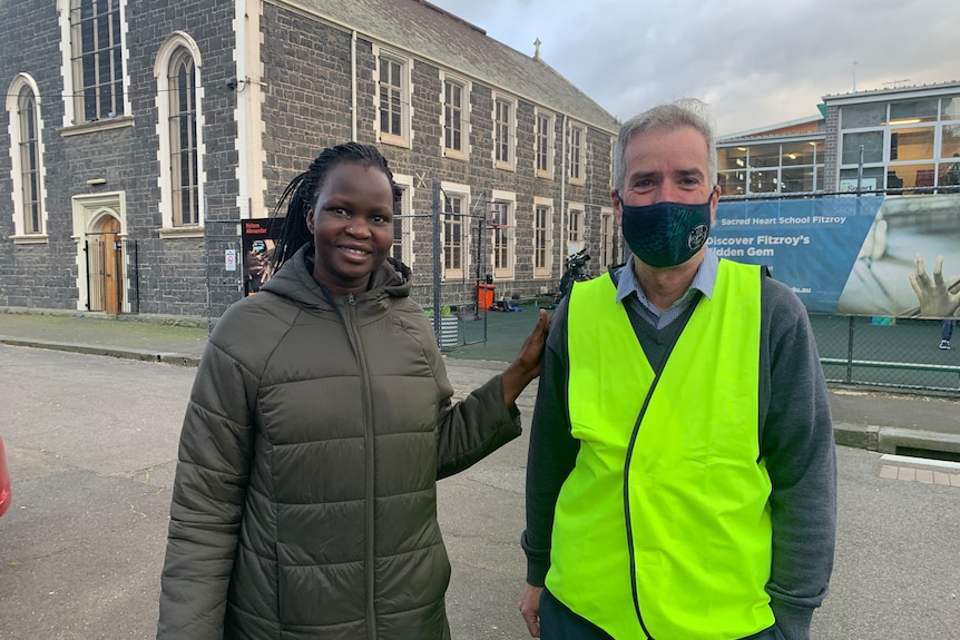 A grey-haired man stands wearing a high-viz vest and a mask in front of a bluestone building besides an African-Australian woman