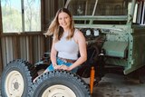 A young woman sits in front of an old vehicle with exposed engine, in front of her are two tyres