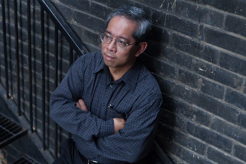 Man of Asian decent wearing navy shirt and glasses standing against a dark brick wall