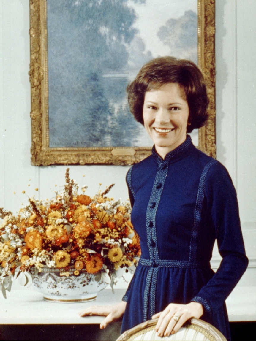 Ritratto di una donna sorridente in piedi con un vestito davanti ad alcuni fiori in un vaso e un dipinto incorniciato sul muro.