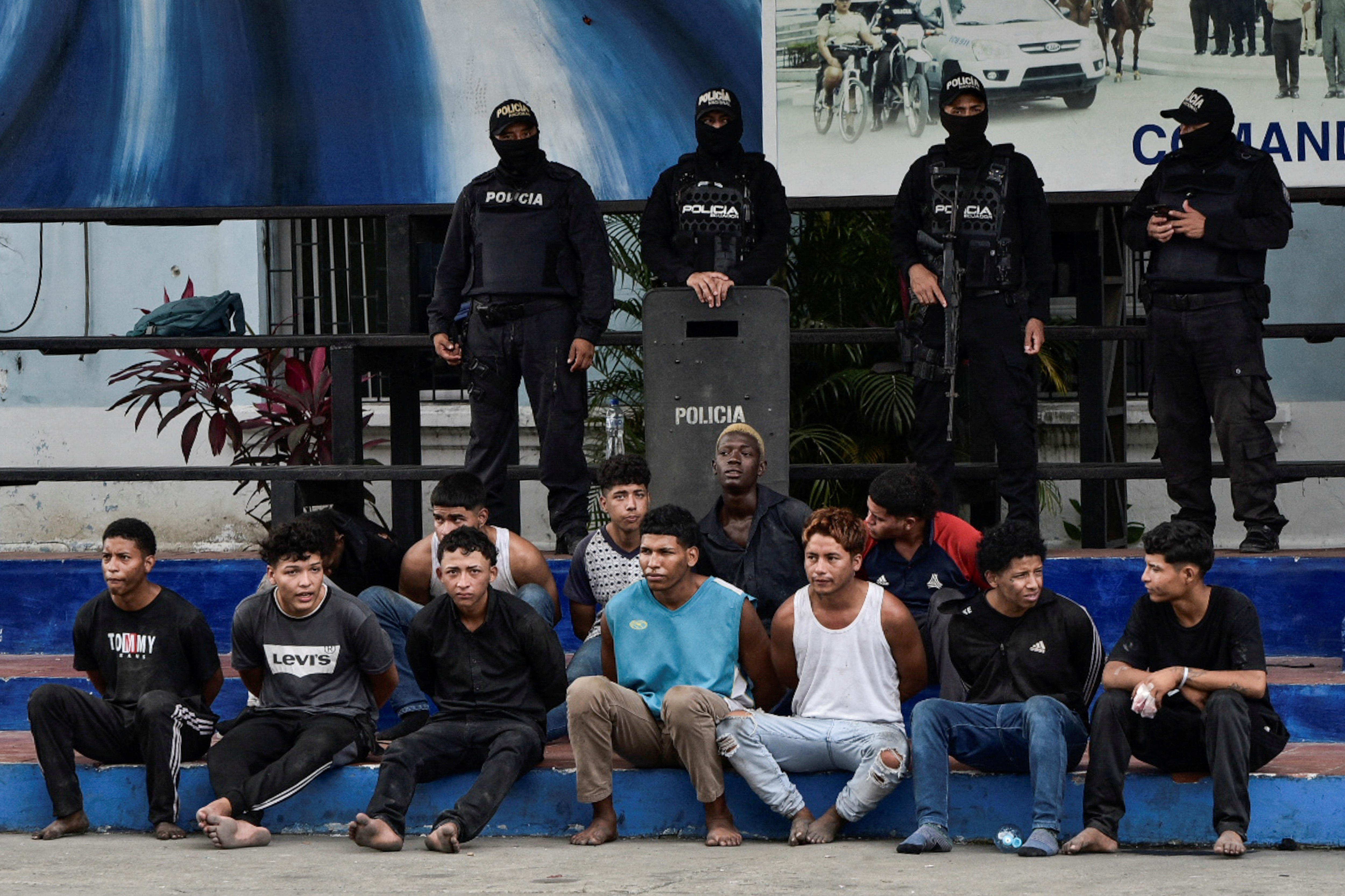 Four police officers stand behind twelve men seated on a set of stairs.